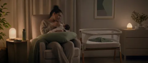 Calm nighttime nursery with a tired parent, feeding setup, and nearby bassinet for safer newborn sleep.