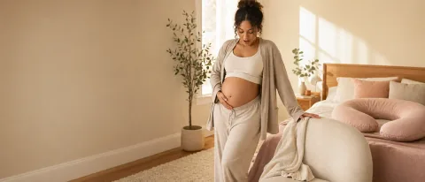 Pregnant woman pausing calmly beside a chair in a warm bedroom while supporting her lower belly.